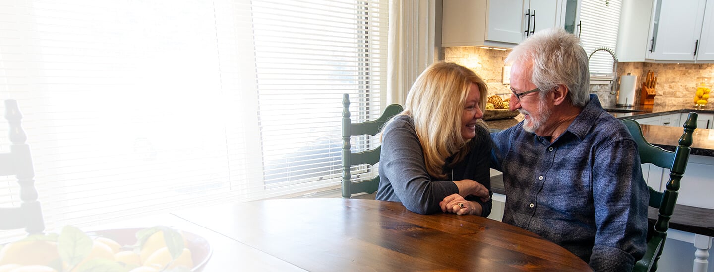A smiling couple sitting at a kitchen table, holding hands and engaging in a warm conversation.